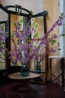 Красивые свежеобрезанные длинные ветки с зелеными листьями и фиолетовыми цветами, стоящими в стеклянной банке с водой на круглом деревянном столе в старой потрепанной комнате с зеркалами — стоковое фото