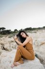Stylish young woman sitting on rocks of shoreline, touching hair and looking in camera — Stock Photo