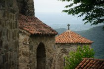 Medieval stone building with red tiled roof and Christian cross on top standing nearby old architectural ensemble in same style with overgrown with trees mountains and valleys on background - foto de stock