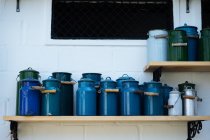 Blaue und grüne Gartenwasserkanister aus Metall mit Deckeln und Holzbügeln in verschiedenen Größen stehen in Reihen auf hellen Holzregalen an weißer Ziegelwand — Stockfoto