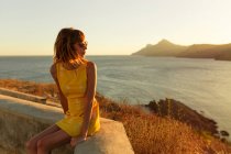 Mulher em vestido curto elegante e óculos de sol sentado no terraço com vista incrível da paisagem marinha ao pôr do sol — Fotografia de Stock