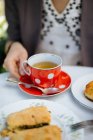 Female hand holding red ceramic polka-dotted mug of tea on saucer on garden table — Stock Photo