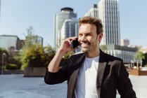 Усміхнений елегантний чоловік посміхається говорити по телефону, стоячи на вулиці сучасного міста в сонячний день — стокове фото