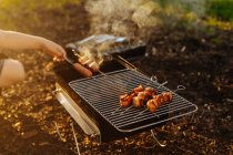 Human hand preparing bacon and sausages on skewers grilling on burning charcoal in portable griddle outdoors — Stock Photo