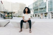Улыбающаяся афроамериканка, сидящая на городской улице и держащая смартфон — стоковое фото