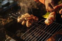 Mãos masculinas preparando bacon e salsichas em espetos grelhando na queima de carvão em grelha portátil ao ar livre — Fotografia de Stock