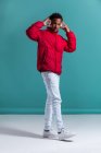 Hombre de moda en chaqueta hinchada roja y auriculares de pie sobre fondo azul - foto de stock