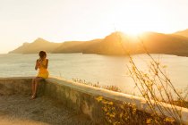 Женщина в стильном коротком платье и солнечных очках, сидящая на террасе с удивительным видом на море на закате — стоковое фото