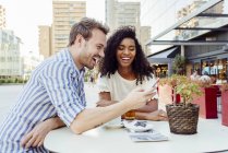 Симпатичная мультирасовая пара, улыбающаяся и просматривающая современный смартфон, сидя за столом в открытом кафе вместе — стоковое фото