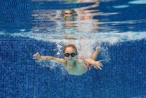 Мальчик в очках плавает в голубом бассейне под водой с воздушными пузырьками — стоковое фото