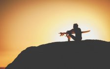 Вид збоку темна фігура чоловічого серфера, що сидить на пагорбі і тримає дошку в руках на задньому освітленому фоні на заході сонця — стокове фото