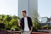 Elegant guy using smartphone while leaning on railing on street of modern city — Stock Photo