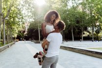 Мужчина поднимает смеющуюся женщину, стоя на аллее парка в солнечный день — стоковое фото
