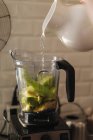 Versare l'acqua dalla brocca di ceramica nel bicchiere di plastica del frullatore riempito con miscela di frutta e verdura per frullato — Foto stock