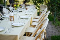 Beautiful table with light cloth and rattan chairs around set with empty wine glasses and plates outdoors on pavement with trees and grass around — Stock Photo