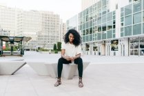 Smiling African-American woman sitting on city street and holding smartphone — Stock Photo
