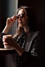 Элегантная женщина в солнцезащитных очках и с платком, держа бумажную чашку кофе и опираясь на стол — стоковое фото