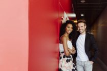 Bonito homem apoiando-se na parede vermelha e flertando com a mulher afro-americana no corredor de construção — Fotografia de Stock