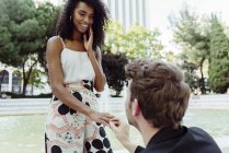 Caucásico hombre poniendo anillo de compromiso en el dedo de la mujer negra mientras propone cerca de la fuente en el parque - foto de stock