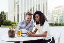 Cute multiracial couple smiling and browsing modern smartphone while sitting at table in outdoor cafe together — Stock Photo