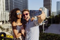 Couple multiracial joyeux posant pour selfie tout en se tenant sur le fond de la ville moderne — Photo de stock