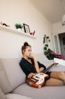 Вдумлива молода жінка з гітарою сидить на дивані вдома — стокове фото