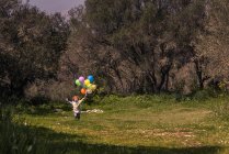 Мальчик-дошкольник бежит по лугу с протянутыми руками с разноцветными воздушными шарами — стоковое фото