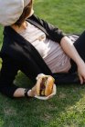 Mulher elegante relaxante na grama no parque e segurando takeaway hambúrguer — Fotografia de Stock