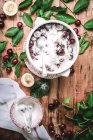 Colocação plana de tigela de cerâmica cheia de açúcar cerejas cobertas com folhas verdes na mesa rústica — Fotografia de Stock