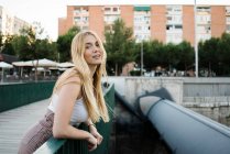 Portrait of attractive young woman smiling and looking at camera while leaning on green railing of bridge over city river — Stock Photo