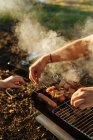 Человеческие руки, готовящие бекон и сосиски на шампурах на гриле на сжигании угля в портативной сковородке на открытом воздухе — стоковое фото