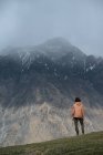 Vista posteriore di maschio in abiti caldi con zaino escursioni in montagna in piedi su erba guardando cresta frastagliata montagna coperta di neve e cime nascoste nelle nuvole — Foto stock