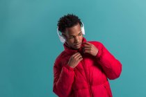 Stylish African American man in headphones and red puffy jacket standing on blue background — Stock Photo