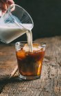Menschliche Hand gießt Milch in eiskalten Brühkaffee auf hölzernem Hintergrund — Stockfoto