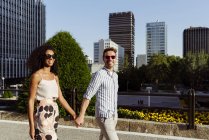 Счастливая многорасовая пара, улыбающаяся и держащаяся за руки во время прогулки по городской улице вместе в солнечный день — стоковое фото