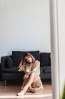 Босоногая женщина в шелковом халате сидит на полу возле дивана и смотрит в камеру — стоковое фото