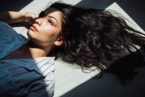 Молодая задумчивая брюнетка с длинными волосами, лежащая на полу в тени и солнечном свете — стоковое фото