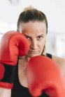 Жінка в боксерських рукавичках стоїть в бойовому положенні і дивиться на камеру під час тренувань — стокове фото
