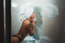 Schuss durch dampfendes Duschglas von nackter Frau in Handtuch auf Kopf und Höschen vor Spiegel — Stockfoto