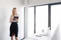 Елегантна жінка в костюмі і використовує планшет, стоячи в яскравому денному світлі всередині сучасного офісу — стокове фото