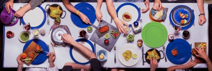 Вид зверху на стіл з різнокольоровими тарілками і людьми, які вечеряють разом, насолоджуючись різними продуктами — стокове фото