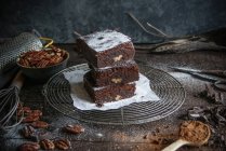 Trozos de delicioso brownie de chocolate en estante de alambre con ingredientes en la superficie de madera oscura - foto de stock