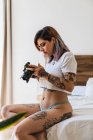Young woman in panties and T-shirt looking at pictures on screen of professional camera on bed — Stock Photo