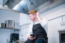 Vista lateral do jovem em cozinheiro uniforme inclinado no balcão da cozinha e usando smartphone moderno enquanto está na cozinha do restaurante — Fotografia de Stock