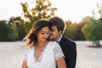 Homem amoroso abraçando noiva bonita em vestido elegante e olhando um para o outro, enquanto em pé na costa arenosa na luz do sol — Fotografia de Stock