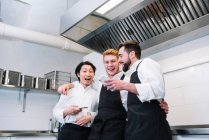 Três homens diversos em uniforme cozinheiro rindo e navegando smartphones enquanto estão de pé na cozinha do restaurante juntos — Fotografia de Stock