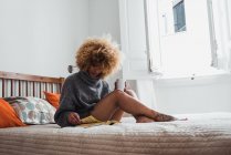 Приваблива етнічна жінка в светрі сидить на ліжку з чашкою кави і читає журнал в денний час — стокове фото