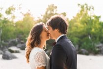 Casal em vestidos de casamento em pé na rocha e abraçando alegremente contra árvores verdes e céu azul — Fotografia de Stock