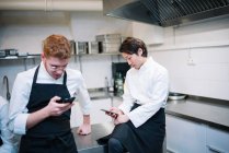 Desde abajo toma de dos chicos en uniforme de cocinero de pie en la cocina del restaurante y navegando por los teléfonos inteligentes durante el descanso - foto de stock