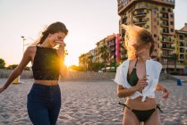 Великолепные энергичные смеющиеся женщины в летней одежде ходят вместе по песку на закате — стоковое фото